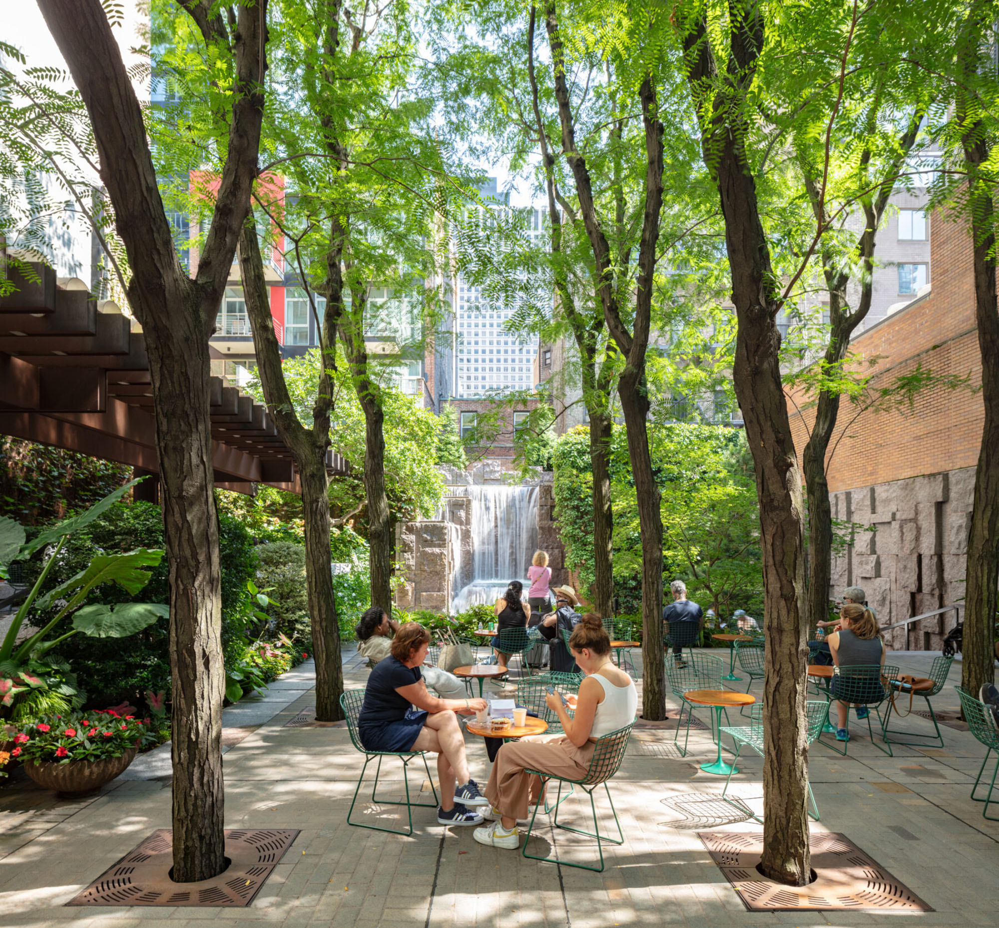 New Yorkers sitting at tables and chairs beneath a lush tree canopy with dappled light and a waterfall in the background